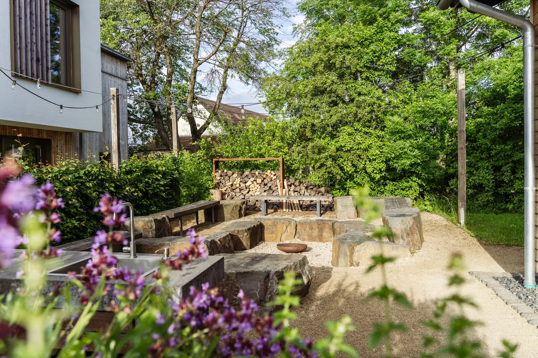 Garten der Waldakademie mit Feuerstelle, Sitzsteinen und Holzstapel
